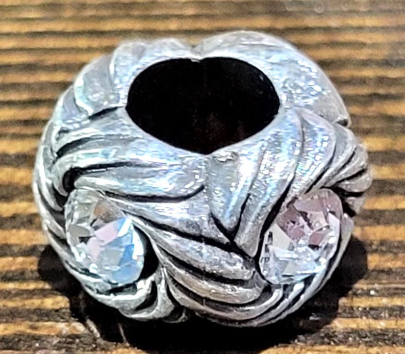 May include: A silver-toned bead with a central hole and swirling, textured design. Two clear, faceted gemstones are set into the bead, one on each side. The bead has a vintage or antique appearance.