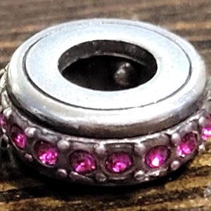 May include: A silver-toned charm with a circular design. The charm features a band of small, bright pink gemstones around its base. The center of the charm has a large, open hole. The charm is set against a wooden surface.
