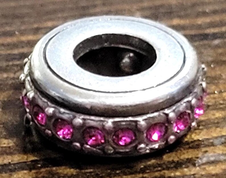May include: A silver-toned charm with a circular design. The charm features a band of small, bright pink gemstones around its base. The center of the charm has a large, open hole. The charm is set against a wooden surface.
