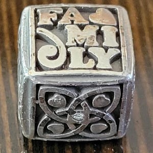 May include: A silver-toned ring with the word "FAMILY" in raised letters. The top of the ring has a gold-colored heart. The sides feature an ornate design with hearts and a small gem.