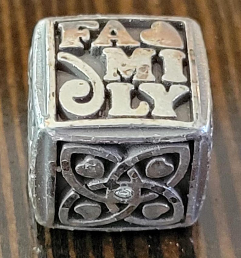 May include: A silver-toned ring with the word "FAMILY" in raised letters. The top of the ring has a gold-colored heart. The sides feature an ornate design with hearts and a small gem.