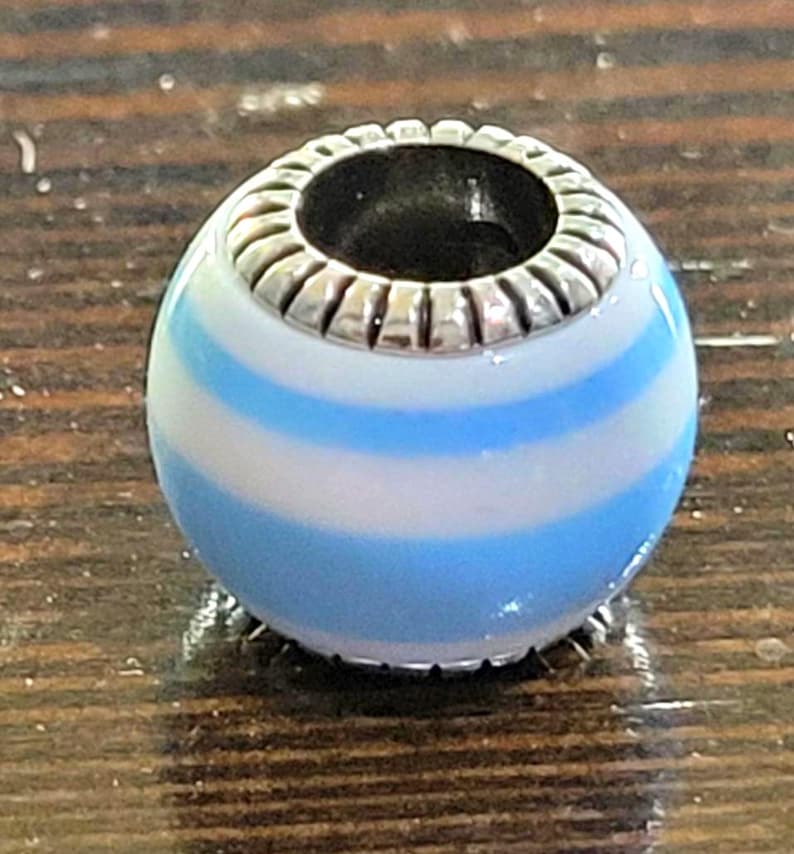 May include: A round bead with a silver-colored top and a blue and white striped design. The bead has a hole through the center and is likely used for jewelry making or as a decorative element.