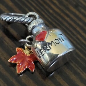 Puede incluir: Un charm de plata con un dise&ntilde;o de botella de jarabe de arce con la palabra "VERMONT" y un coraz&oacute;n rojo. Un charm de hoja de arce roja y dorada cuelga debajo de la botella. El charm tiene un anillo retorcido para sujetarlo.