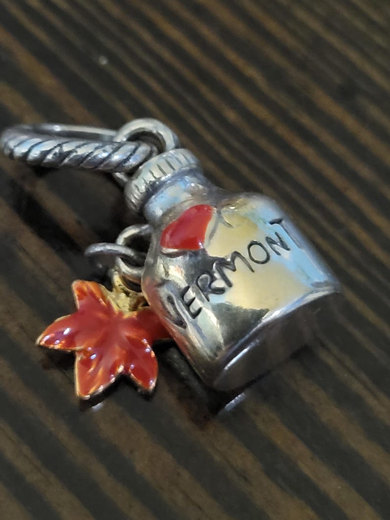 Puede incluir: Un charm de plata con un dise&ntilde;o de botella de jarabe de arce con la palabra "VERMONT" y un coraz&oacute;n rojo. Un charm de hoja de arce roja y dorada cuelga debajo de la botella. El charm tiene un anillo retorcido para sujetarlo.