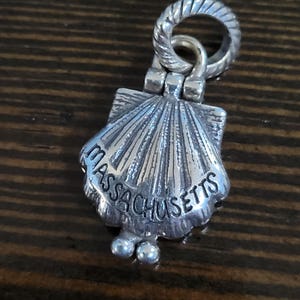 Puede incluir: Un charm plateado con forma de concha de vieira. El charm tiene grabada la palabra "Massachusetts". Tiene un anillo retorcido en la parte superior para sujetarlo y dos peque&ntilde;as esferas en la parte inferior.
