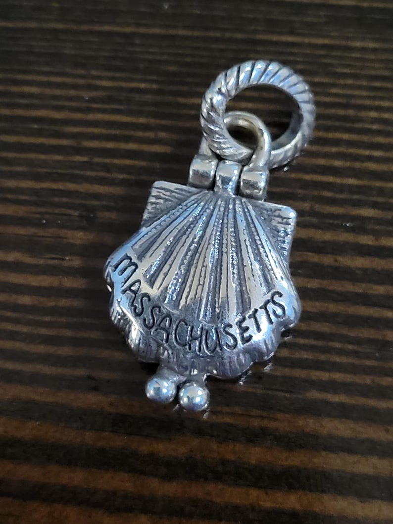 Puede incluir: Un charm plateado con forma de concha de vieira. El charm tiene grabada la palabra "Massachusetts". Tiene un anillo retorcido en la parte superior para sujetarlo y dos peque&ntilde;as esferas en la parte inferior.