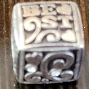 May include: Silver-toned charm with the word "BEST" in raised letters. The charm has decorative swirls, hearts, and a sparkling gem. The charm is a jewelry accessory.