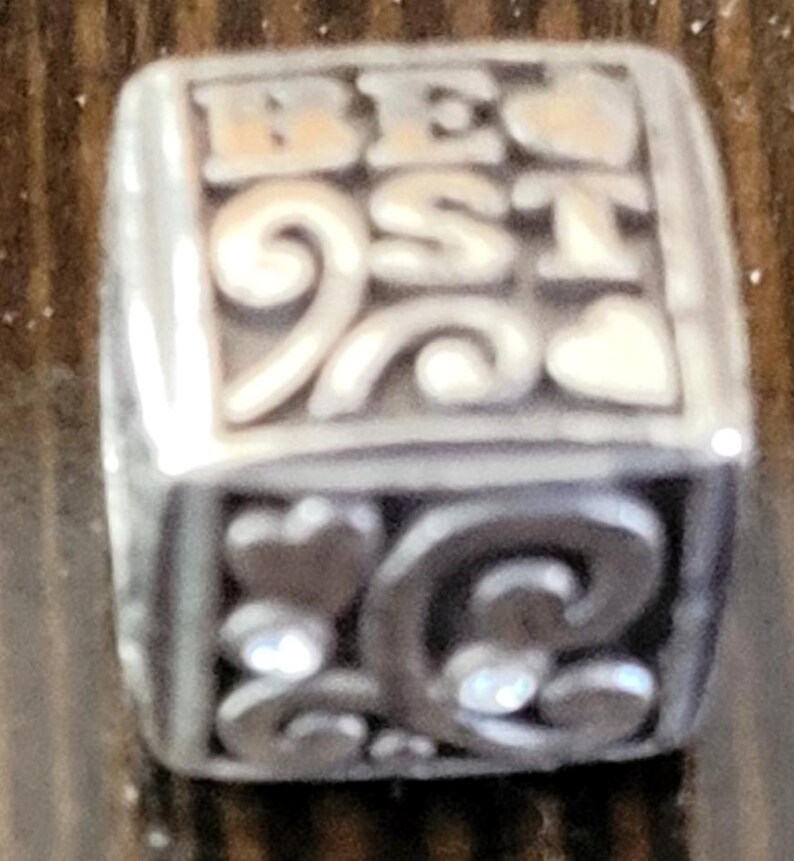 May include: Silver-toned charm with the word "BEST" in raised letters. The charm has decorative swirls, hearts, and a sparkling gem. The charm is a jewelry accessory.
