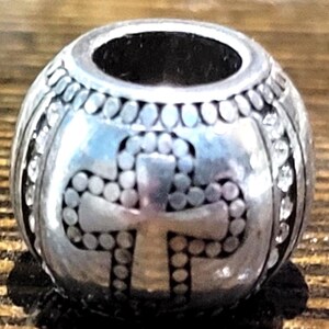 May include: A silver-colored bead with a cross design. The bead has a cylindrical hole and is decorated with small, round accents and clear, sparkling stones. The cross is made of dotted patterns.