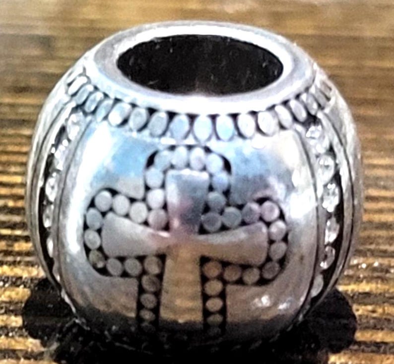 May include: A silver-colored bead with a cross design. The bead has a cylindrical hole and is decorated with small, round accents and clear, sparkling stones. The cross is made of dotted patterns.