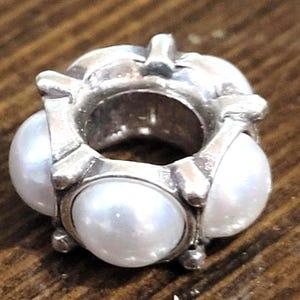 May include: A silver-toned charm with three large, round, white pearls. The charm has a circular design with the pearls set around the outside. The metal has a slightly tarnished appearance, and the pearls have a lustrous sheen. This charm is suitable for bracelets or necklaces.