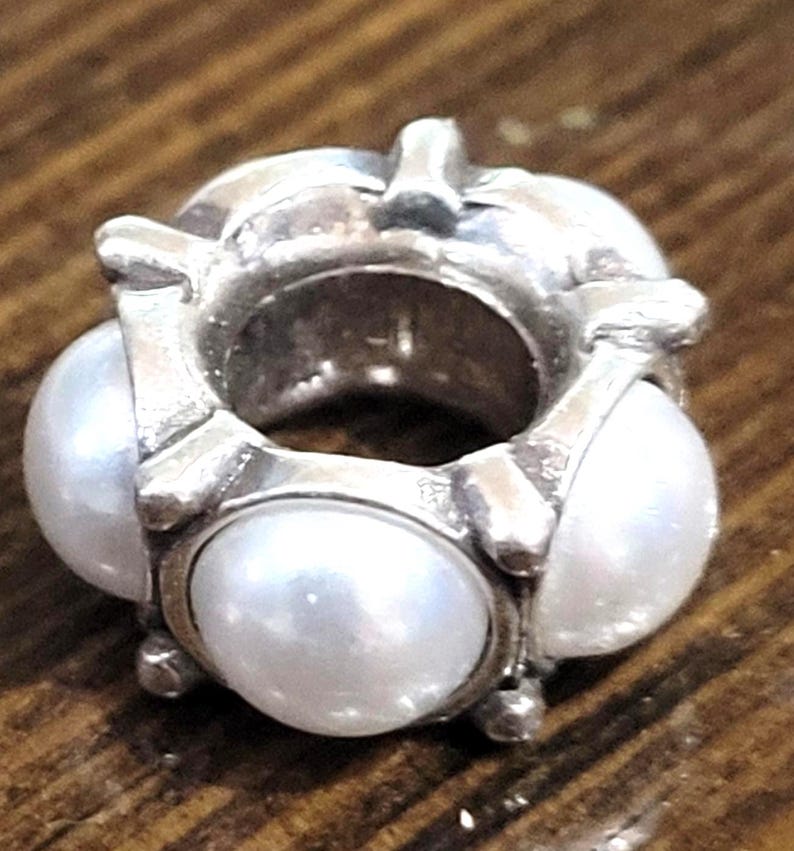 May include: A silver-toned charm with three large, round, white pearls. The charm has a circular design with the pearls set around the outside. The metal has a slightly tarnished appearance, and the pearls have a lustrous sheen. This charm is suitable for bracelets or necklaces.