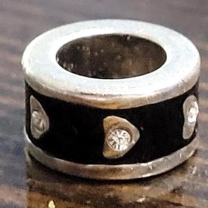 May include: A silver-toned bead with a black band. The band features three silver-colored shapes, each with a clear, sparkling stone. The bead has a circular design with a hole in the center.