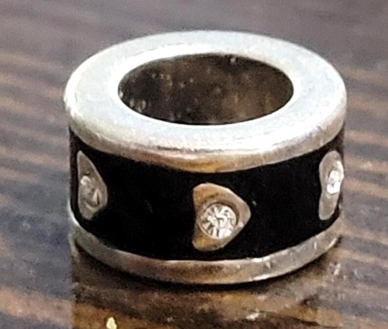 May include: A silver-toned bead with a black band. The band features three silver-colored shapes, each with a clear, sparkling stone. The bead has a circular design with a hole in the center.