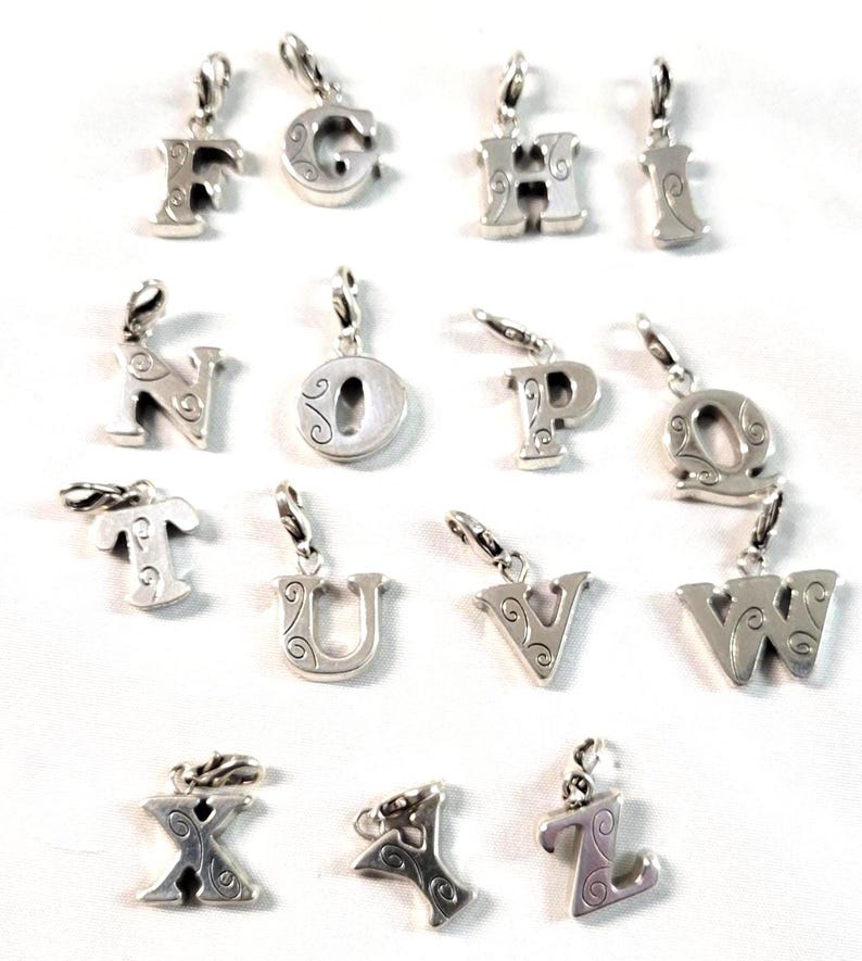 May include: A collection of silver-toned alphabet charms, each with a decorative swirl design. The charms are suspended from small silver loops, and are arranged on a white background. The letters include F, G, H, I, N, O, P, Q, T, U, V, W, X, Y, and Z.
