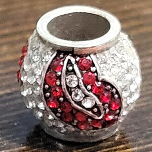 May include: A silver-toned bead with a central design of red and clear crystals. The bead has a cylindrical shape with a wide opening at the top. The design features a pattern of red crystals and clear crystals.