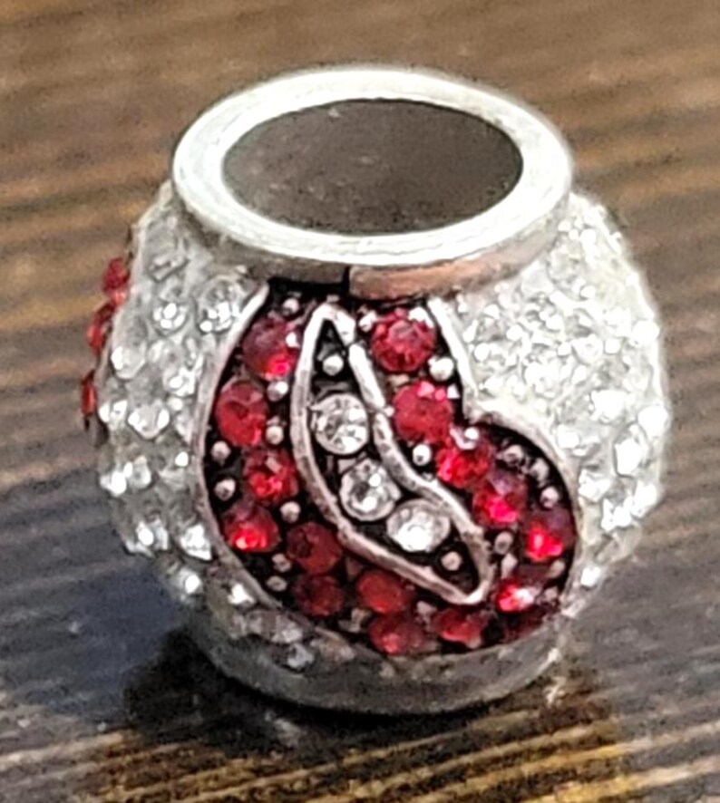 May include: A silver-toned bead with a central design of red and clear crystals. The bead has a cylindrical shape with a wide opening at the top. The design features a pattern of red crystals and clear crystals.