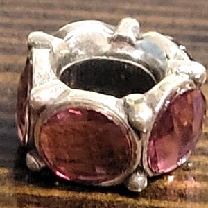 May include: A silver-toned charm with six oval, faceted, pink gemstones. The charm has a cylindrical shape with a large central opening. The gemstones are set into the silver metal, creating a decorative bead.
