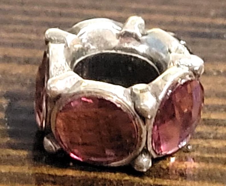 May include: A silver-toned charm with six oval, faceted, pink gemstones. The charm has a cylindrical shape with a large central opening. The gemstones are set into the silver metal, creating a decorative bead.