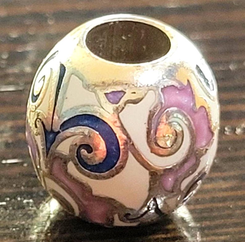 May include: A decorative bead with a silver finish and a central hole. The bead features a cream-colored base with swirling patterns in pink, blue, and gold. The bead is approximately 0.75 inches in diameter.