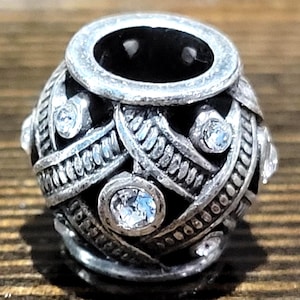 May include: A silver-toned bead with a dark interior and a decorative design. The bead features clear, round gemstones and intricate metalwork. The bead's design includes interwoven patterns and a circular opening at the top.