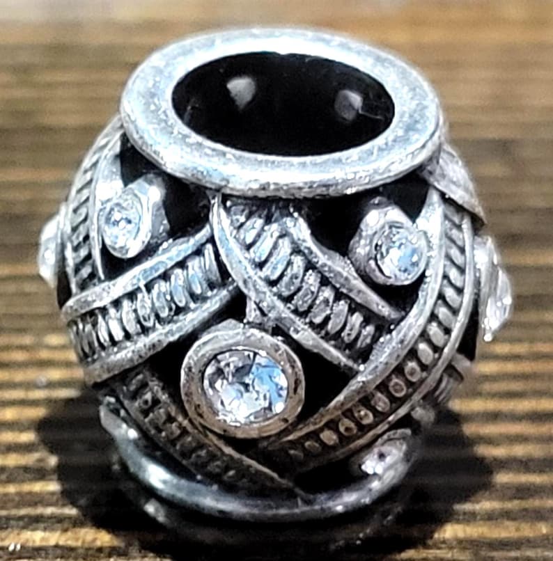 May include: A silver-toned bead with a dark interior and a decorative design. The bead features clear, round gemstones and intricate metalwork. The bead's design includes interwoven patterns and a circular opening at the top.