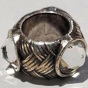 May include: A silver-toned ring with a woven band design and three clear, faceted gemstones. The ring has a vintage or antique appearance, with a slightly tarnished finish. The gemstones are set in a bezel setting.