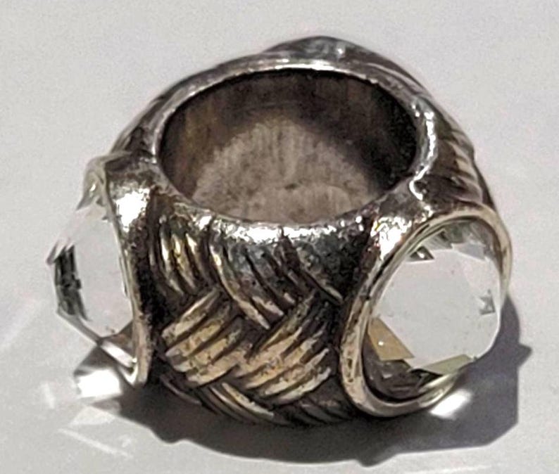 May include: A silver-toned ring with a woven band design and three clear, faceted gemstones. The ring has a vintage or antique appearance, with a slightly tarnished finish. The gemstones are set in a bezel setting.