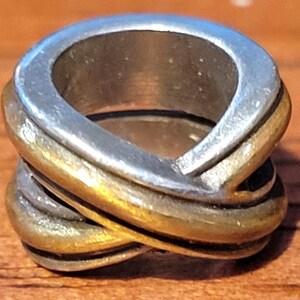 May include: A two-tone ring with a silver top and gold-colored band. The ring has an interwoven design, with the bands crossing over each other. The ring is made of metal and has a polished finish. The ring is a piece of jewelry.