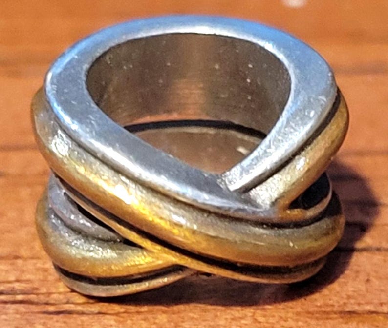 May include: A two-tone ring with a silver top and gold-colored band. The ring has an interwoven design, with the bands crossing over each other. The ring is made of metal and has a polished finish. The ring is a piece of jewelry.