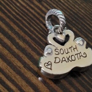 Puede incluir: Un colgante de plata con las palabras "SOUTH DAKOTA" grabadas. El colgante tiene un coraz&oacute;n y un peque&ntilde;o s&iacute;mbolo de coraz&oacute;n. El dije est&aacute; sobre una superficie de madera.