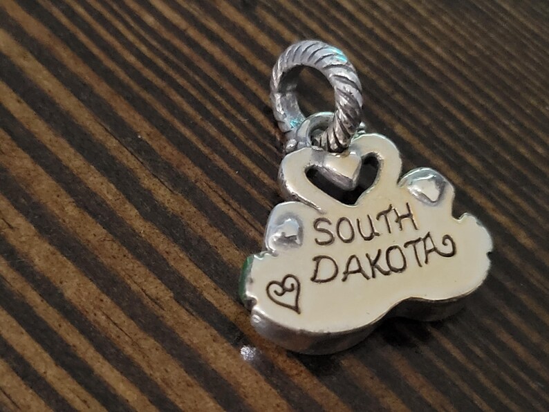 Puede incluir: Un colgante de plata con las palabras "SOUTH DAKOTA" grabadas. El colgante tiene un coraz&oacute;n y un peque&ntilde;o s&iacute;mbolo de coraz&oacute;n. El dije est&aacute; sobre una superficie de madera.