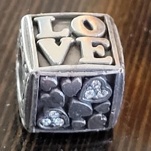 May include: Silver-toned charm with the word "LOVE" in raised letters. The charm has a cube shape with heart and spade designs, some with clear crystal accents. A romantic-themed jewelry piece.
