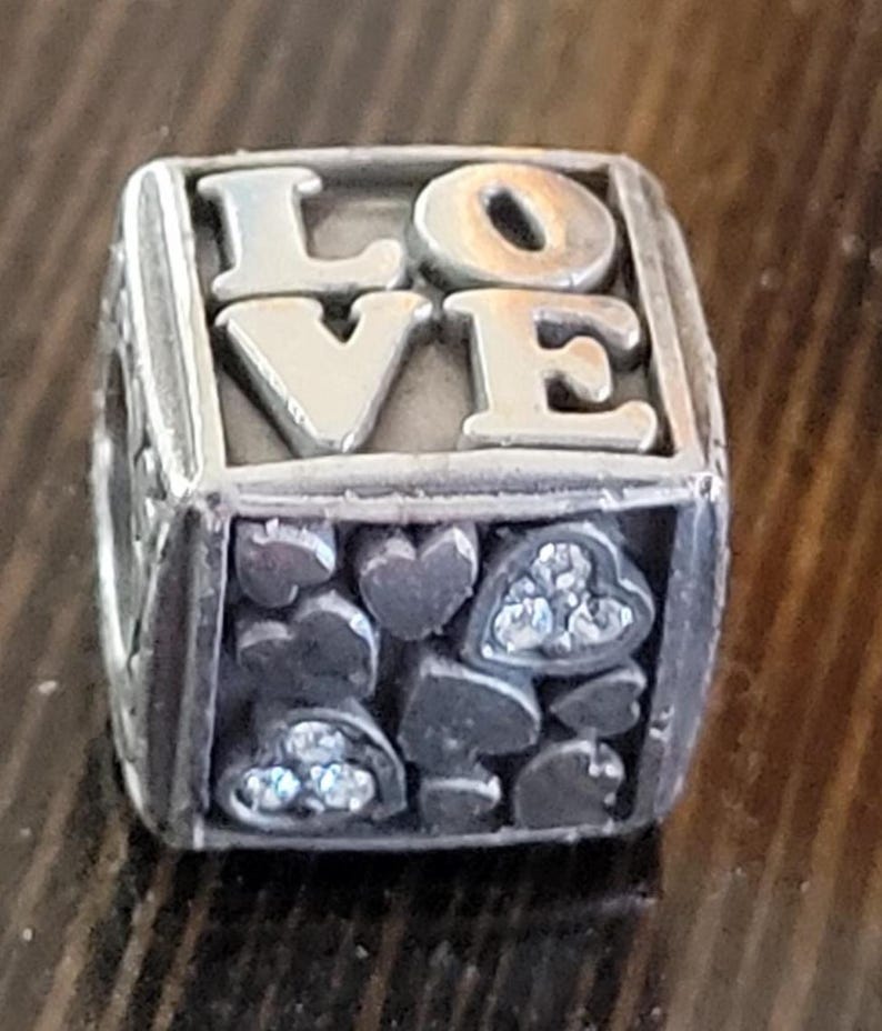 May include: Silver-toned charm with the word "LOVE" in raised letters. The charm has a cube shape with heart and spade designs, some with clear crystal accents. A romantic-themed jewelry piece.