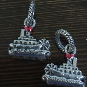 Puede incluir: Dos colgantes plateados con forma de barcos fluviales. Cada colgante tiene una rueda, m&uacute;ltiples cubiertas y las palabras "RIVER BOAT" y "MISSISSIPPI" grabadas. Un peque&ntilde;o coraz&oacute;n rojo est&aacute; unido a la parte superior de cada colgante.