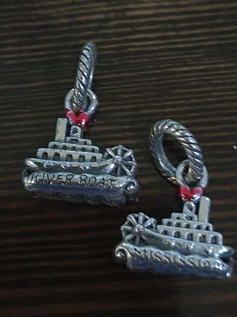 Puede incluir: Dos colgantes plateados con forma de barcos fluviales. Cada colgante tiene una rueda, m&uacute;ltiples cubiertas y las palabras "RIVER BOAT" y "MISSISSIPPI" grabadas. Un peque&ntilde;o coraz&oacute;n rojo est&aacute; unido a la parte superior de cada colgante.