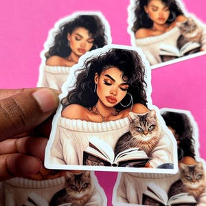 Op de afbeelding: Een sticker met een vrouw met lang, krullend bruin haar die een boek leest terwijl ze een bruin-witte kat vasthoudt. De vrouw draagt een witte trui en heeft een gouden ketting om. De sticker is op een roze achtergrond.