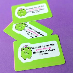 May include: Three green rectangular stickers with rounded corners are stacked on a purple surface. Each sticker features a cartoon frog and the text "Excited for all the good things that are in store for me."