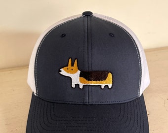 May include: A navy blue and white trucker hat with a white mesh back. The hat features an embroidered image of a corgi dog.