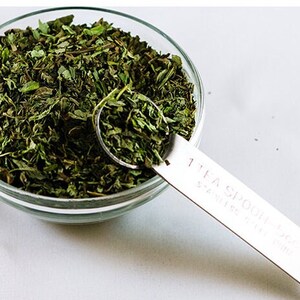May include: A glass bowl filled with dried green herbs, with a 1 teaspoon measuring spoon filled with the herbs.