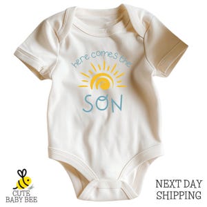A white baby onesie with a sun graphic and the text "here comes the SON" printed on it.