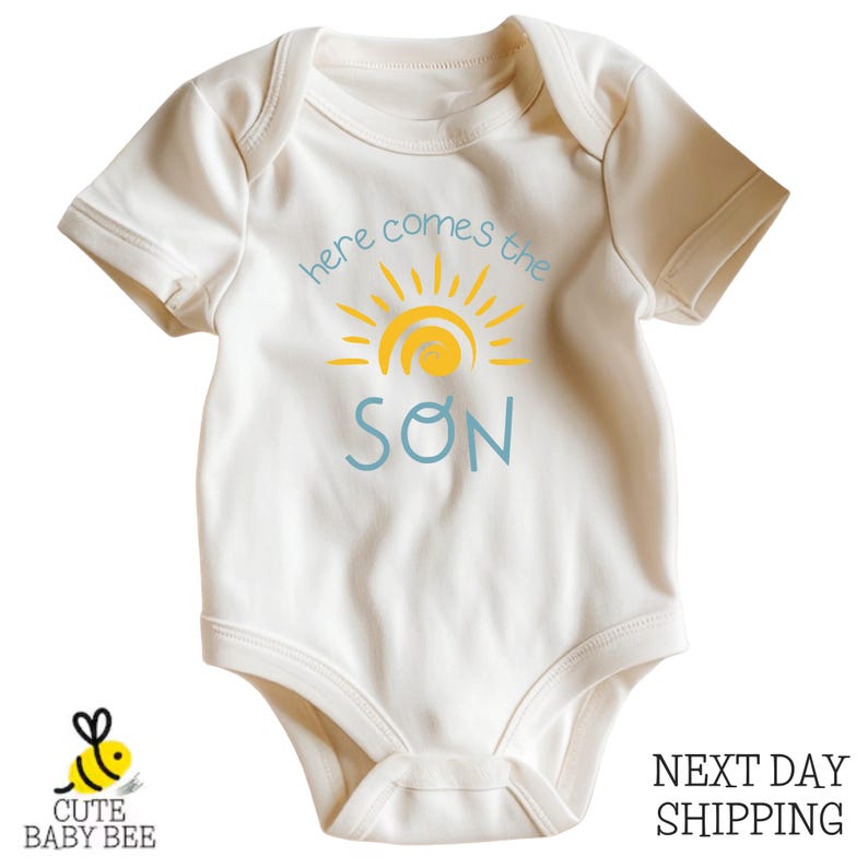 A white baby onesie with a sun graphic and the text "here comes the SON" printed on it.