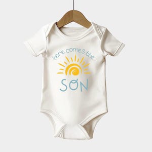 a baby's bodysuit with the words here comes the son on it