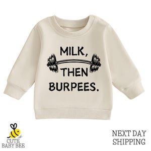 May include: Cream-colored baby sweatshirt with the text "MILK, THEN BURPEES" in black, along with a barbell graphic. The sweatshirt has long sleeves and a crew neck. The logo "CUTE BABY BEE" is in the bottom left corner.