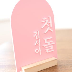 Puede incluir: Un letrero de acr&iacute;lico rosa con texto coreano blanco, sostenido por una base de madera natural. El letrero tiene una parte superior arqueada y est&aacute; dise&ntilde;ado para colocarse de pie. El texto est&aacute; en una fuente estilizada.