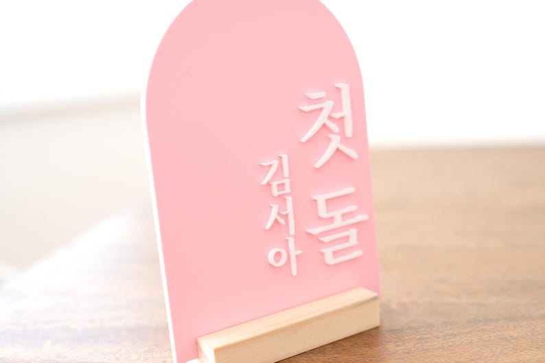Puede incluir: Un letrero de acr&iacute;lico rosa con texto coreano blanco, sostenido por una base de madera natural. El letrero tiene una parte superior arqueada y est&aacute; dise&ntilde;ado para colocarse de pie. El texto est&aacute; en una fuente estilizada.