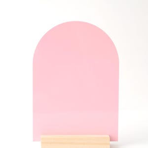 Puede incluir: Un letrero rosa en forma de arco con una base de madera clara. El letrero es de color rosa liso y tiene una parte superior redondeada. La base es un bloque rectangular de madera natural.