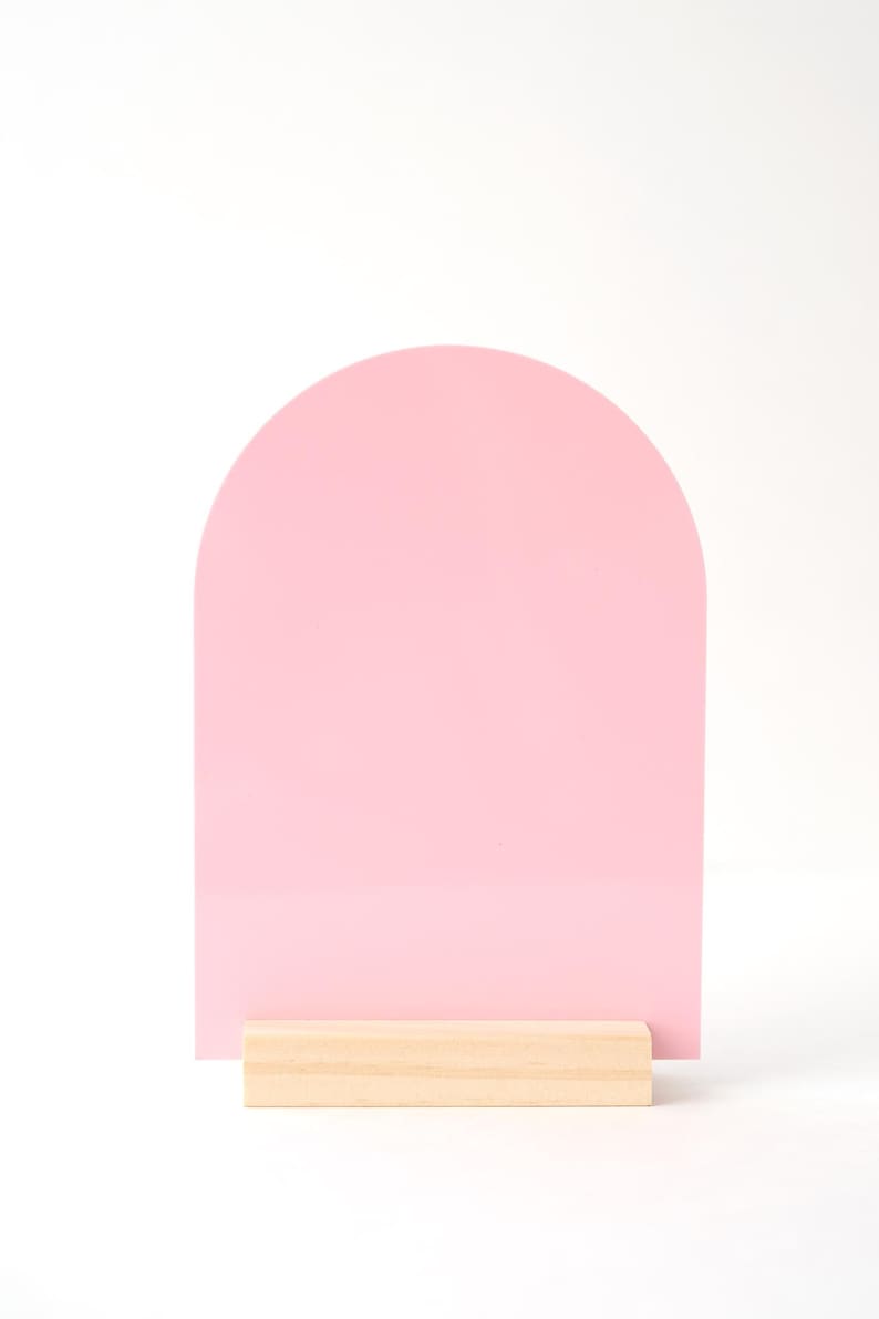 Puede incluir: Un letrero rosa en forma de arco con una base de madera clara. El letrero es de color rosa liso y tiene una parte superior redondeada. La base es un bloque rectangular de madera natural.