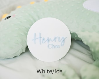 May include: A white, round sign with the name "Henry Choi" in light blue script. The sign is set against a soft, mint green textured background. The text "White/Ice" is at the bottom.