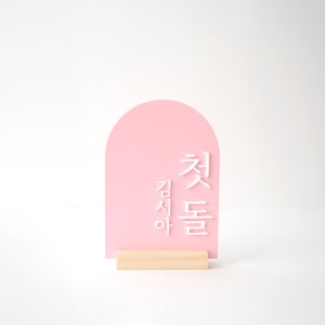 Puede incluir: Un letrero de acr&iacute;lico rosa con una parte superior arqueada y texto coreano blanco. El letrero est&aacute; montado sobre una peque&ntilde;a base de madera rectangular. El fondo es blanco.